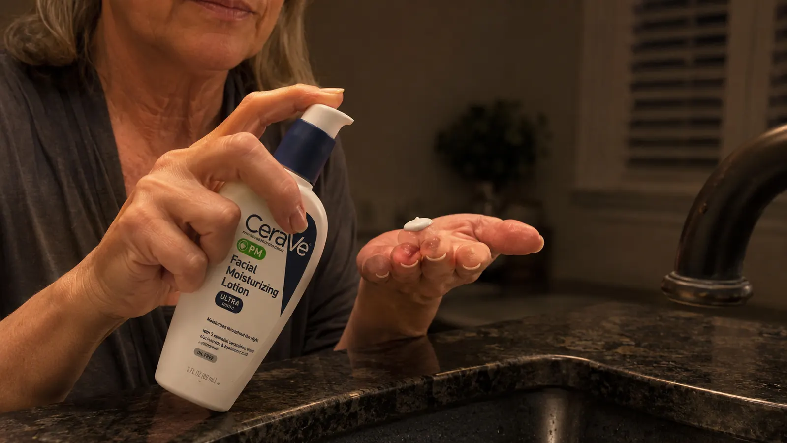 A woman's hand pressing a pump of lotion onto her fingertips over a bathroom sink at night