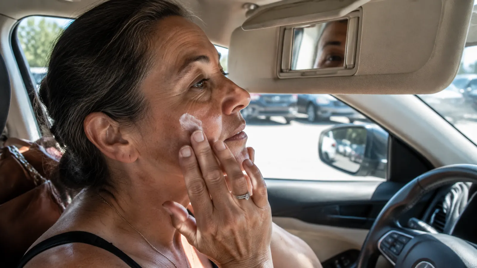 A woman in her fifties reapplies sunscreen from a small travel-size bottle in a car with the sun visor mirror down, midday light, a purse and sunglasses visible on the passenger seat