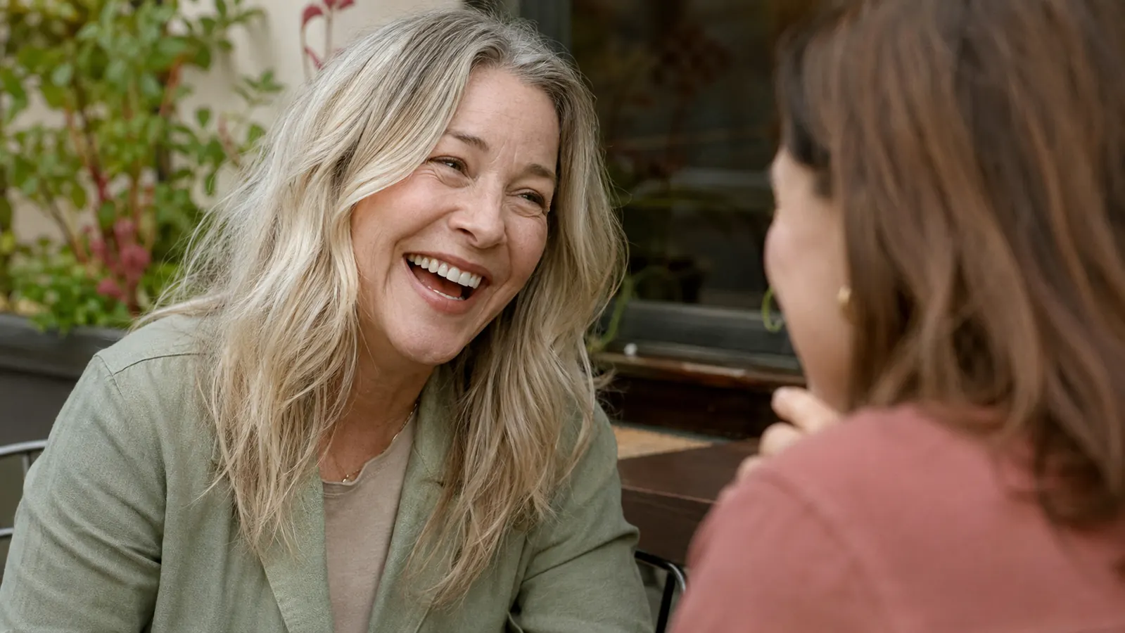 A woman in her fifties laughing candidly with a friend at an outdoor cafe table, her under-eye area visible and unconcealed