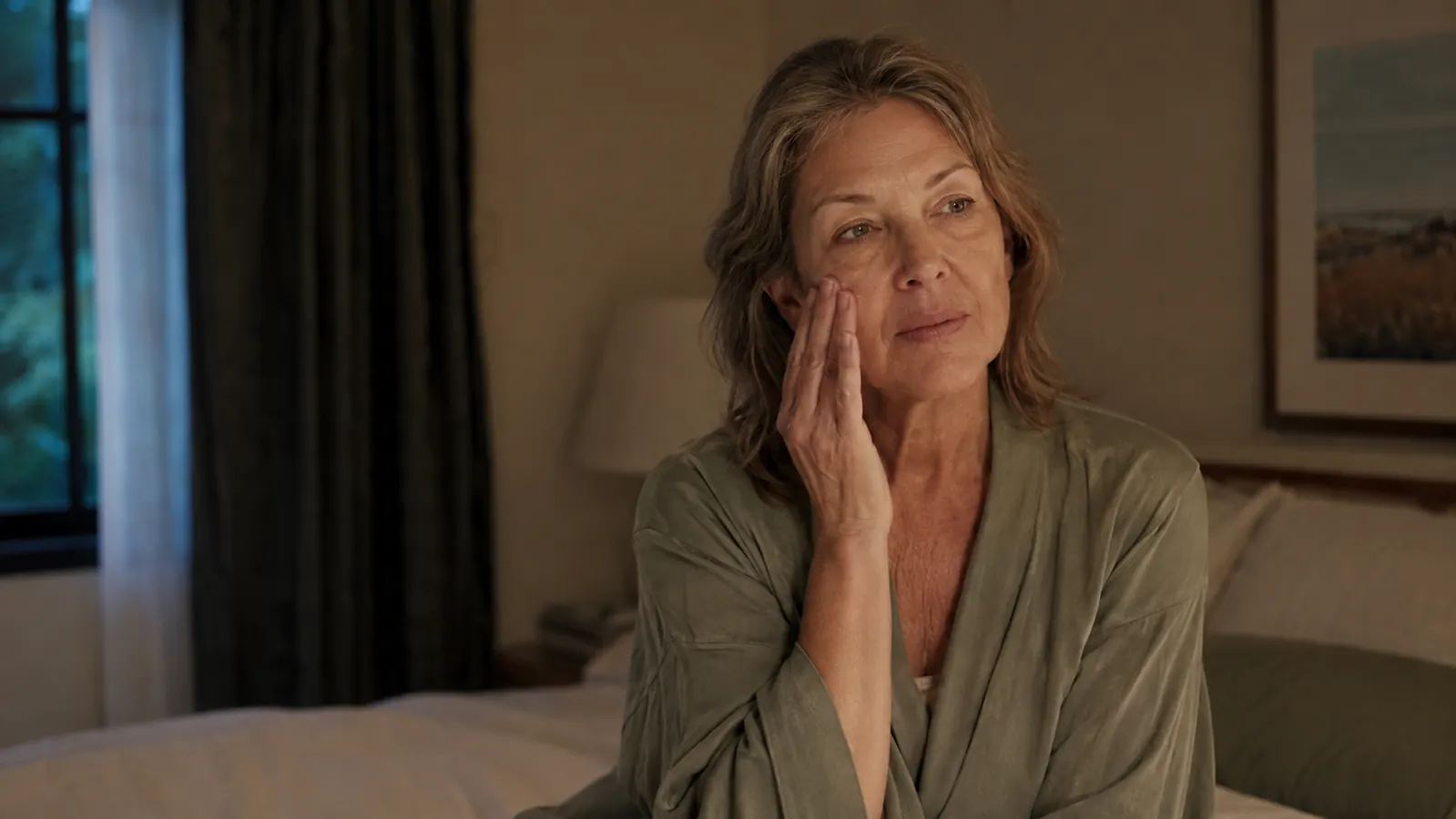 A woman in her fifties sitting on the edge of a bed applying moisturizer to her cheek in dim evening light