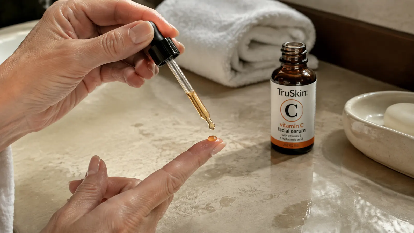 Close-up of a hand tilting the glass dropper of a vitamin C serum bottle to apply a drop onto a fingertip, over a bathroom sink with a folded white towel nearby