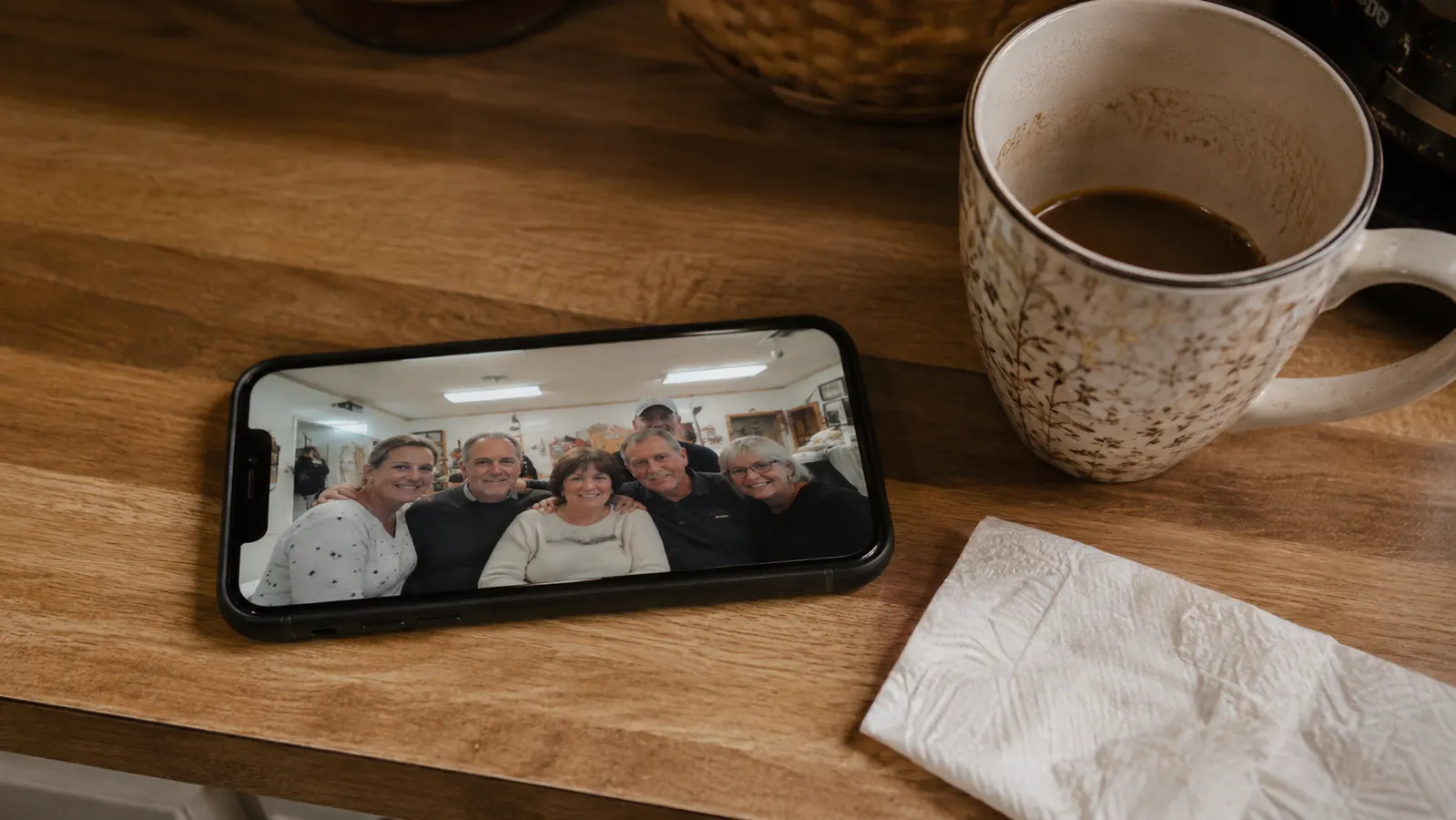 A phone propped on a kitchen counter showing a blurry group photo from a retirement lunch, next to a coffee mug